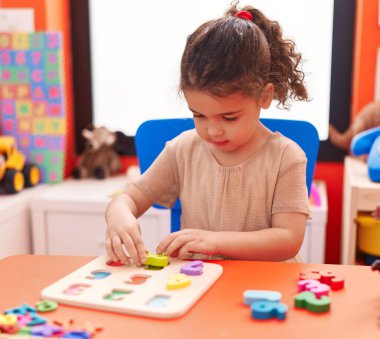 Adorable hispanic girl playing with maths puzzle game sitting on table at kindergarten