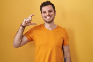 Young hispanic man standing over yellow background smiling and confident gesturing with hand doing small size sign with fingers looking and the camera. measure concept. 