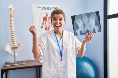 Young woman holding pelvis radiography celebrating victory with happy smile and winner expression with raised hands 