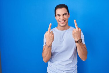Young hispanic man standing over blue background showing middle finger doing fuck you bad expression, provocation and rude attitude. screaming excited 