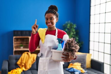 Young african american with braids cleaning the house holding cleaning products surprised with an idea or question pointing finger with happy face, number one 