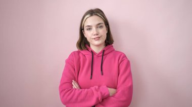 Young blonde woman standing with relaxed expression and arms crossed gesture over isolated pink background