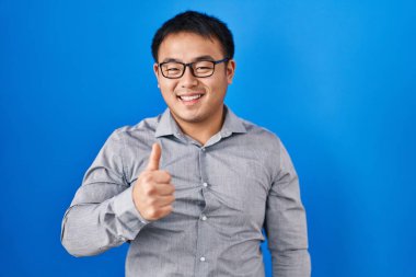 Young chinese man standing over blue background doing happy thumbs up gesture with hand. approving expression looking at the camera showing success. 