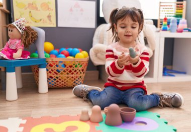 Adorable hispanic girl playing with toys sitting on floor at kindergarten