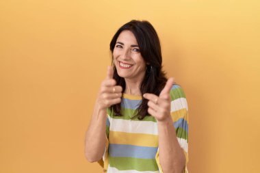 Middle age hispanic woman standing over yellow background pointing fingers to camera with happy and funny face. good energy and vibes. 