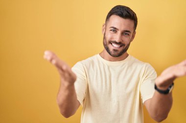 Handsome hispanic man standing over yellow background smiling cheerful offering hands giving assistance and acceptance. 