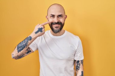 Sarı arka planda dikilen İspanyol dövmeli genç bir adam. El ve burunla işaret ediyor. Neşeli gülümsüyor. güzellik konsepti 