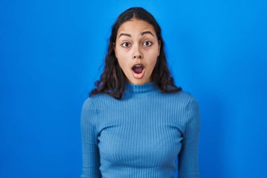 Young brazilian woman standing over blue isolated background afraid and shocked with surprise and amazed expression, fear and excited face. 
