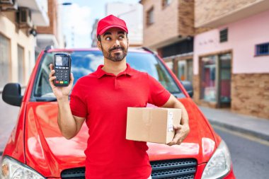 Young hispanic man with beard wearing delivery uniform and cap holding dataphone smiling looking to the side and staring away thinking. 