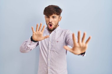 Arab man with beard standing over blue background doing stop gesture with hands palms, angry and frustration expression 