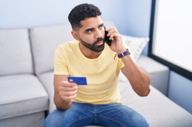 Young arab man talking on smartphone and credit card sitting on sofa at home