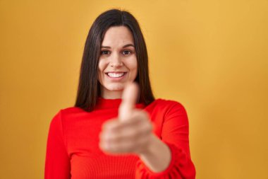 Young hispanic woman standing over yellow background smiling friendly offering handshake as greeting and welcoming. successful business. 