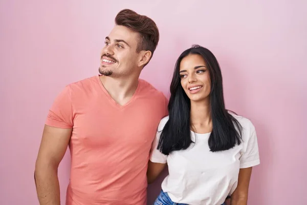 若いヒスパニック系のカップルピンクの背景に離れて顔に笑顔で側に見て上に立って 自然な表現 自信を持って — ストック写真