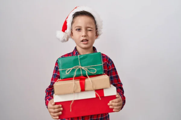 小さなヒスパニック男の子身に着けているクリスマスの帽子保持プレゼントウィンク見ますでザ カメラとともにセクシー表現 陽気で幸せな顔 — ストック写真