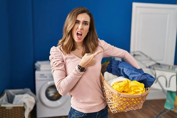 Young woman holding laundry basket pointing aside worried and nervous with forefinger, concerned and surprised expression