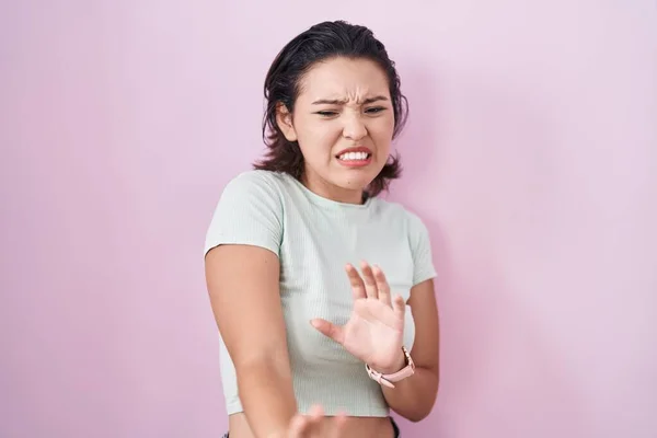 Hispanic young woman standing over pink background disgusted expression, displeased and fearful doing disgust face because aversion reaction.