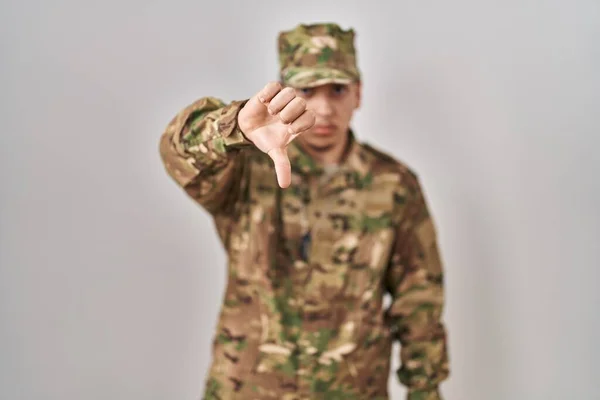 Young arab man wearing camouflage army uniform looking unhappy and angry showing rejection and negative with thumbs down gesture. bad expression.