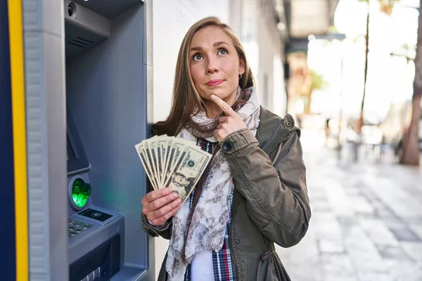 Молодая Блондинка Банкнотами Руках Банкомата Серьезное Лицо Думающая Вопросе Рукой — стоковое фото