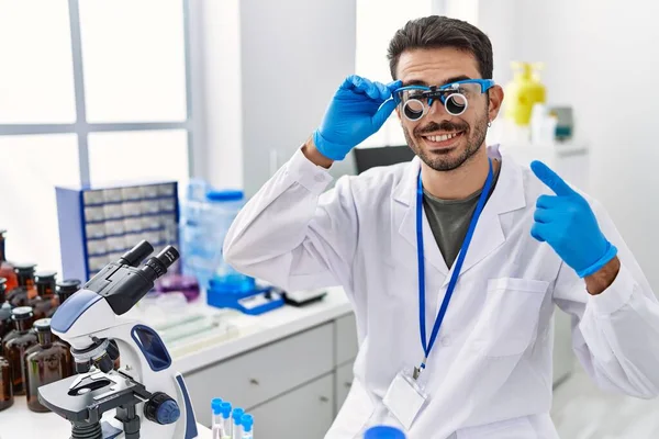 Young hispanic man working at scientist laboratory wearing magnifying glasses smiling happy pointing with hand and finger