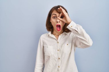 Young beautiful woman standing casual over blue background doing ok gesture shocked with surprised face, eye looking through fingers. unbelieving expression. 