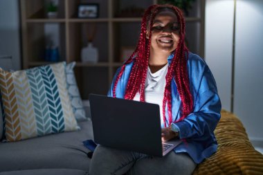 African american woman with braided hair using computer laptop at night looking away to side with smile on face, natural expression. laughing confident. 