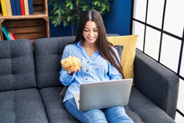 Young beautiful hispanic woman watching movie eating chips potatoes at home