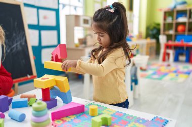 Adorable hispanic girl playing with construction blocks standing at kindergarten