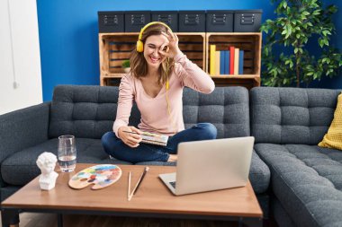 Young beautiful woman practicing painting looking at online tutorial smiling happy doing ok sign with hand on eye looking through fingers 