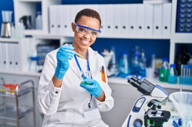African american woman scientist smiling confident pouring liquid on test tube at laboratory