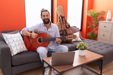 Sakallı İspanyol adam evde klasik gitar çalıyor mutlu ve pozitif gülümsüyor, başparmağıyla mükemmel ve onay işareti yapıyor. 