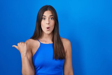 Hispanic woman standing over blue background surprised pointing with hand finger to the side, open mouth amazed expression. 