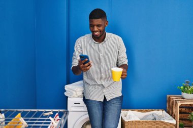 Young african american man using smartphone and drinking coffee waiting for washing machine at laundry room