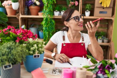 Young beautiful hispanic woman florist talking on smartphone sitting on table at flower shop