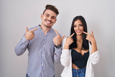 Young hispanic couple standing over white background smiling cheerful showing and pointing with fingers teeth and mouth. dental health concept. 