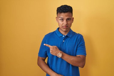 Young hispanic man standing over yellow background pointing aside worried and nervous with forefinger, concerned and surprised expression 