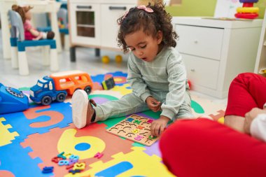 Adorable hispanic girl playing with maths puzzle game sitting on floor at kindergarten