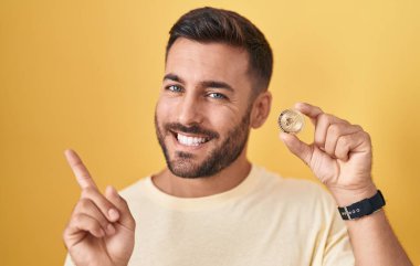 Handsome hispanic man holding uniswap cryptocurrency coin smiling happy pointing with hand and finger to the side 