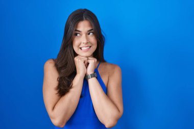 Hispanic woman standing over blue background laughing nervous and excited with hands on chin looking to the side 