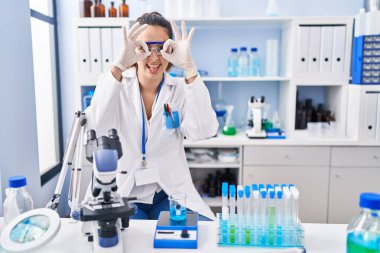 Young hispanic woman working at scientist laboratory doing ok gesture like binoculars sticking tongue out, eyes looking through fingers. crazy expression. 