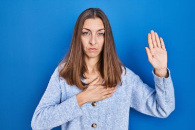 Young woman standing over blue background swearing with hand on chest and open palm, making a loyalty promise oath 