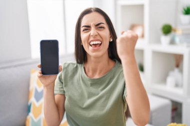 Young brunette woman holding smartphone showing blank screen annoyed and frustrated shouting with anger, yelling crazy with anger and hand raised 