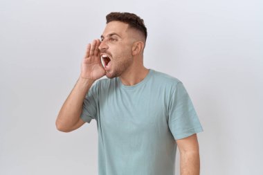 Hispanic man with beard standing over white background shouting and screaming loud to side with hand on mouth. communication concept. 