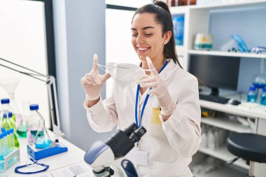 Young beautiful hispanic woman scientist smiling confident holding medical mask at laboratory