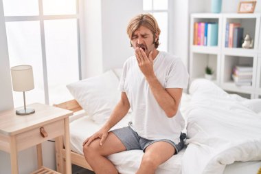 Young blond man sitting on bed yawning at bedroom