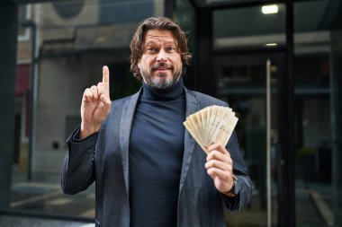 Elinde 100 Danimarka kronu tutan yakışıklı orta yaşlı bir adam bir fikir veya soru işareti ile gülümsüyor. 