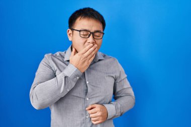 Young chinese man standing over blue background bored yawning tired covering mouth with hand. restless and sleepiness. 