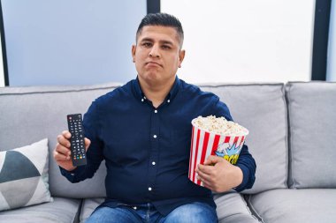 Hispanic young man eating popcorn using tv control relaxed with serious expression on face. simple and natural looking at the camera. 