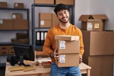 Hispanic man with beard working at small business ecommerce holding packages winking looking at the camera with sexy expression, cheerful and happy face. 