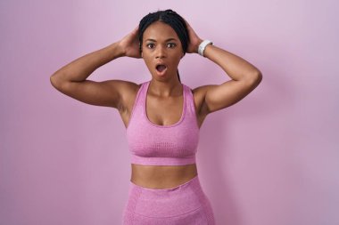 Pembe arka planda spor kıyafetleri giyen Afro-Amerikalı kadın çıldırmış ve elleri başının üstünde korkmuş, ağzı açık şok geçirmekten korkmuş ve şaşırmış. 