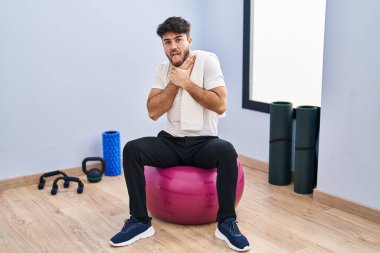 Hispanic man with beard sitting on pilate balls at yoga room shouting and suffocate because painful strangle. health problem. asphyxiate and suicide concept. 